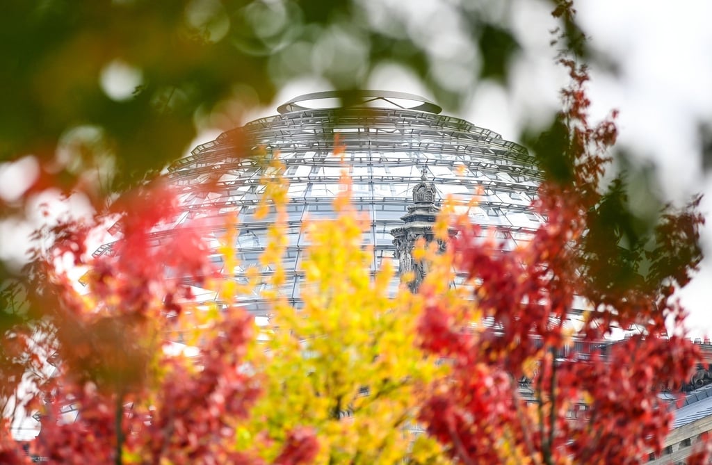 Rot-gelb-grün herbstlich verfärbte Blätter sind an Bäumen vor der Reichstagskuppel zu sehen.