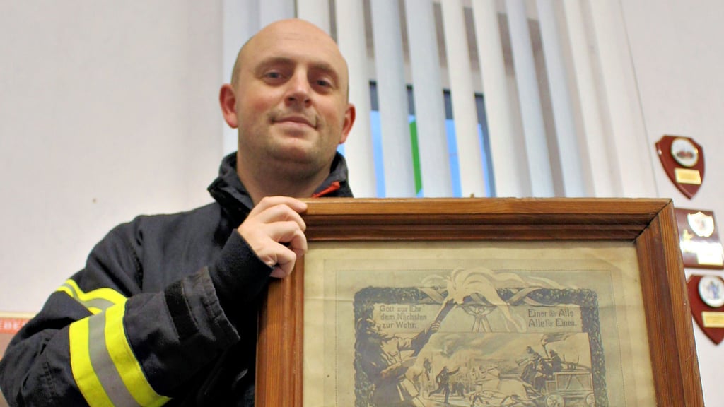 Ortswehrleiter Sascha Schmidt präsentiert ein Zeitdokument, das noch heute hinter Glas im Feuerwehrhaus hängt. Es dokumentiert, dass 1931 das 50-jährige Bestehen gefeiert wurde.