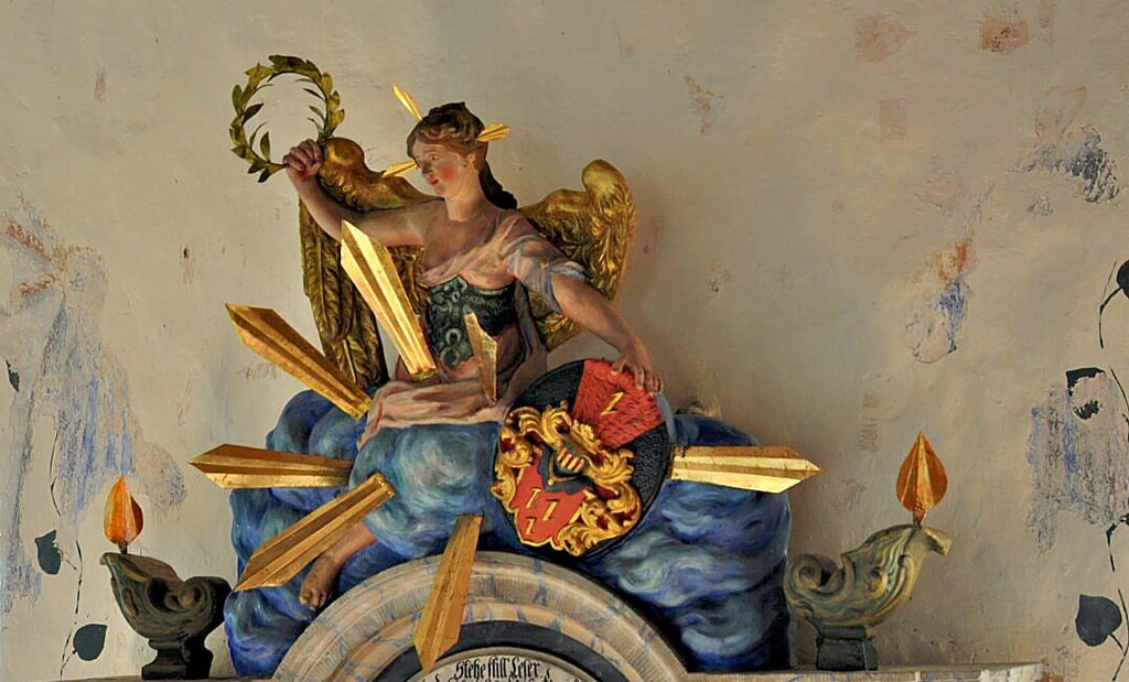 Die weibliche und geflügelte Friedensfigur ganz oben auf dem Epitaph ist wieder komplett mit Strahlen- und Efeukranz. Auch von den beiden Öllampen war vorher nur eine zu sehen. Die andere war abgebrochen.