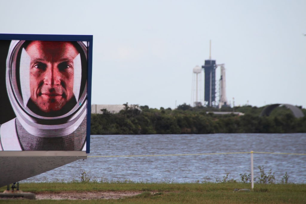 Matthias Maurer auf einem Video-Bildschirm vor dem Launchpad 39A in Cape Canaveral, an dem schon die Falcon-9-Rakete mit Crew Dragon-Kapsel aufgestellt ist.