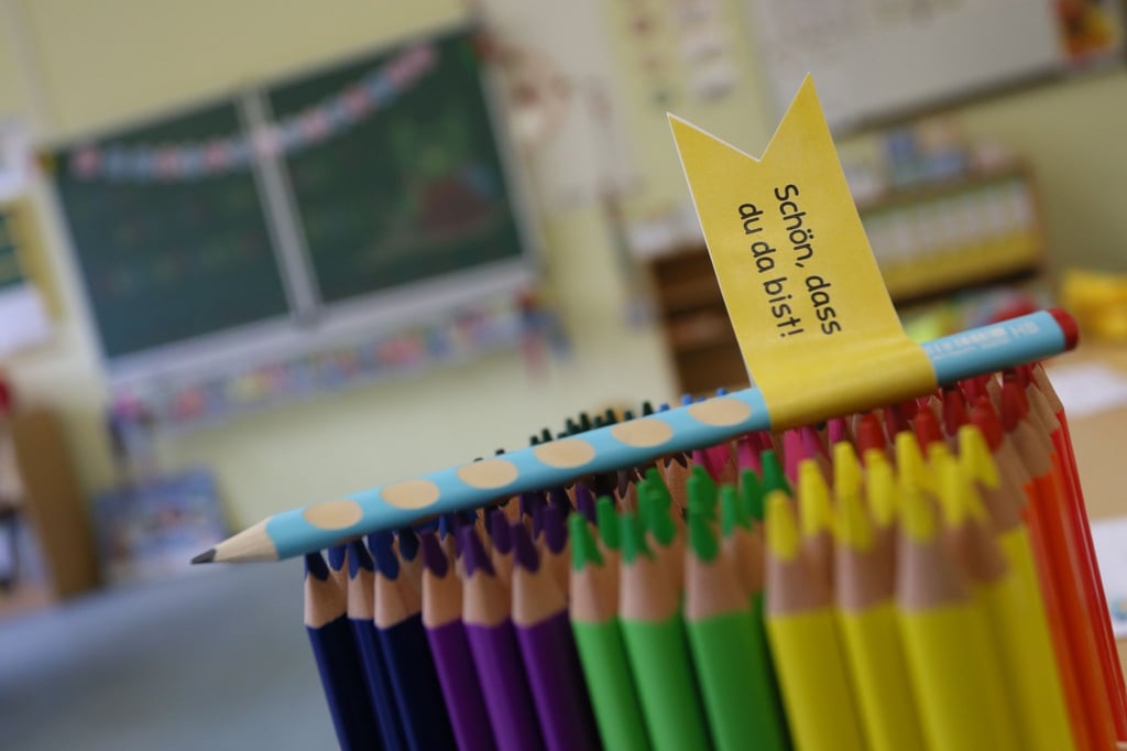 Ein Farbstift mit der Aufschrift „Schön das du da bist“ steckt in einem Klassenraum zwischen vielen bunten Stiften.