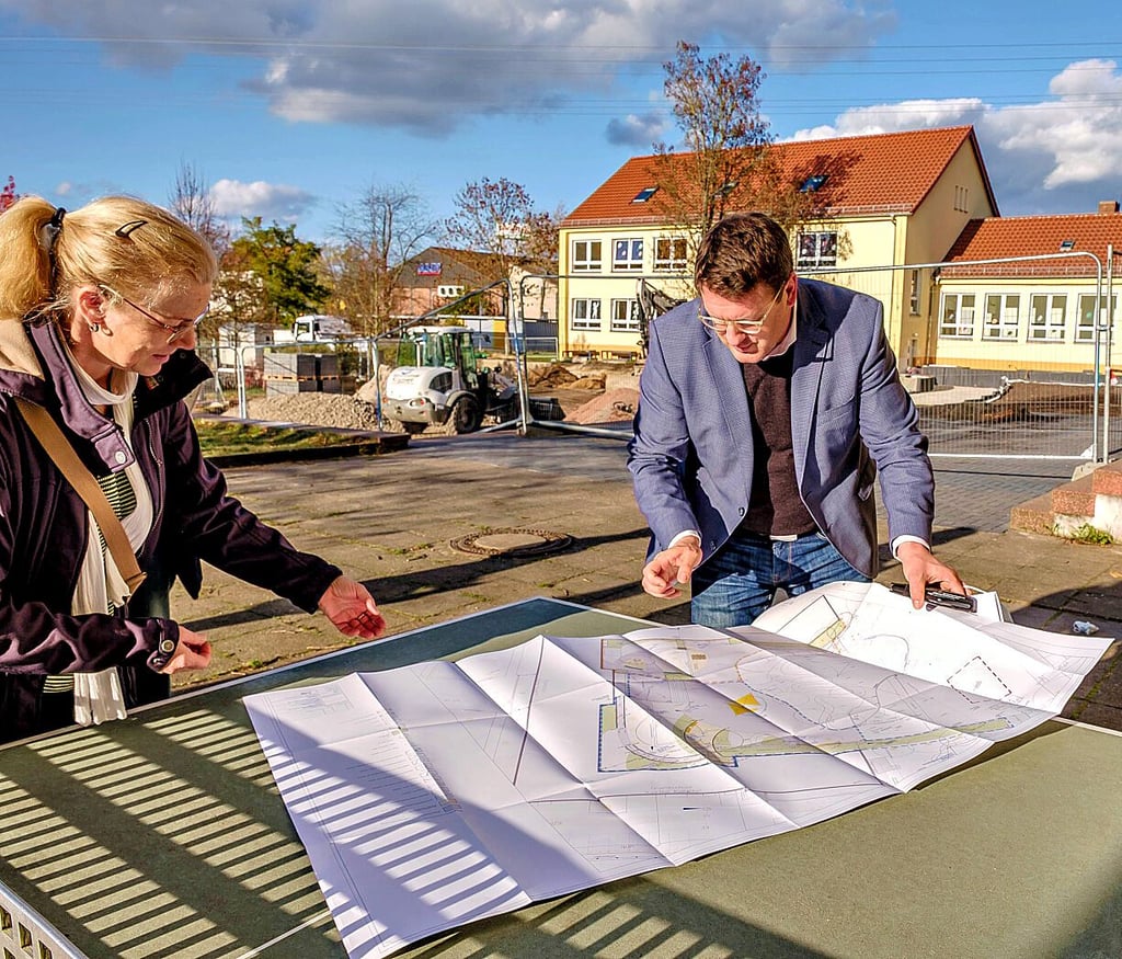 Bauleiterin Ina Heyer und Oberbürgermeister Torsten Zugehör zeigen den Plan für den neuen Schulhof, im Hintergrund die Baustelle.