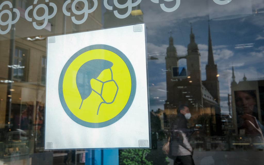 Ein Schild mit einer Maske am Kaufhof in Halle am Marktplatz weist auf das Tragen von Masern im Geschäft hin. Daneben spiegeln sich die Hausmanstürme.