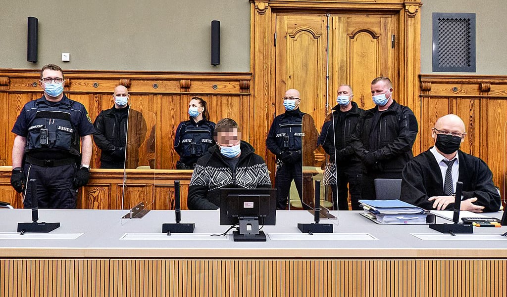 Der Angeklagte ist an Händen und Füßen gefesselt und wird von sechs Justizbeamten bewacht.