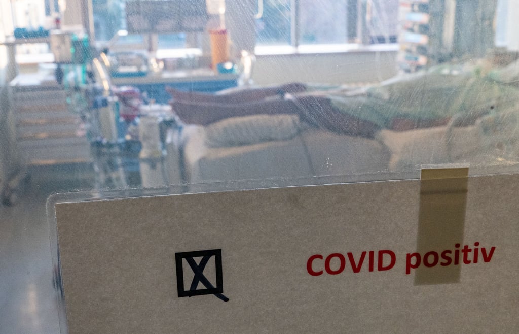 Ein schwer an Covid-19 erkrankter Patient liegt hinter einem Schild mit dem Hinweis "Covid positiv" in einem Isolierzimmer auf einer Corona-Intensivstation.