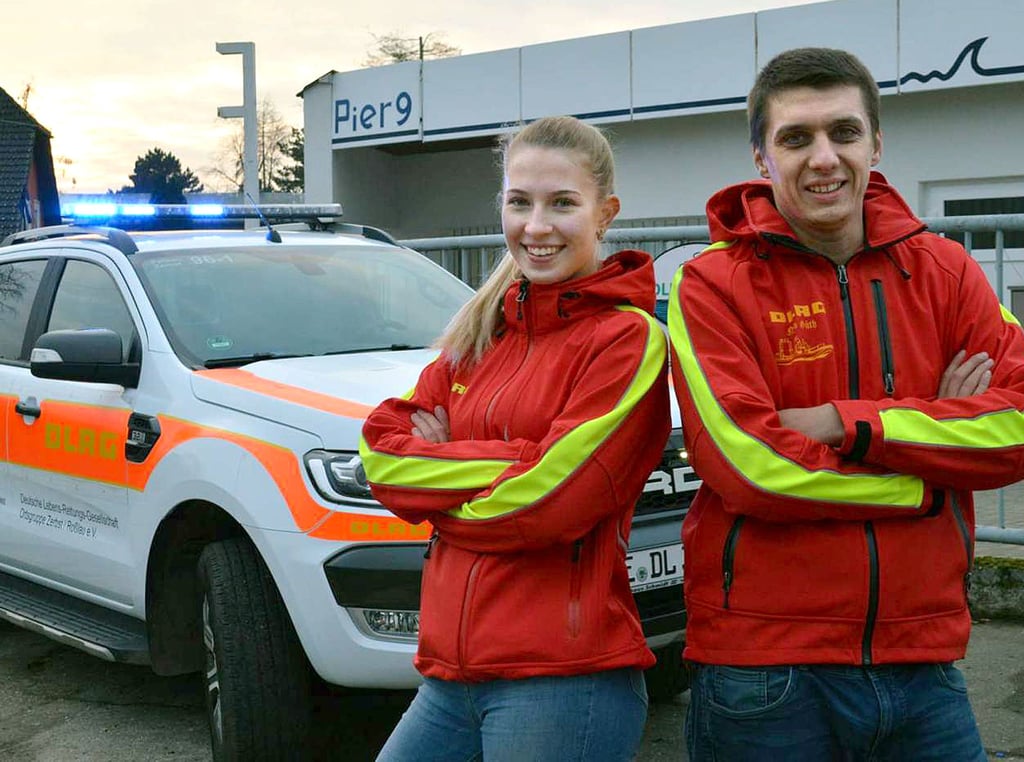 Sophia Volik und Nico Güth von der Ortsgruppe Zerbst/Anhalt der DLRG vor einem der Einsatzfahrzeuge. Im Hintergrund das mit Einegmitteln und Spenden ausgebaute und in diesem Jahr eingeweihte Jugendheim Pier 9.