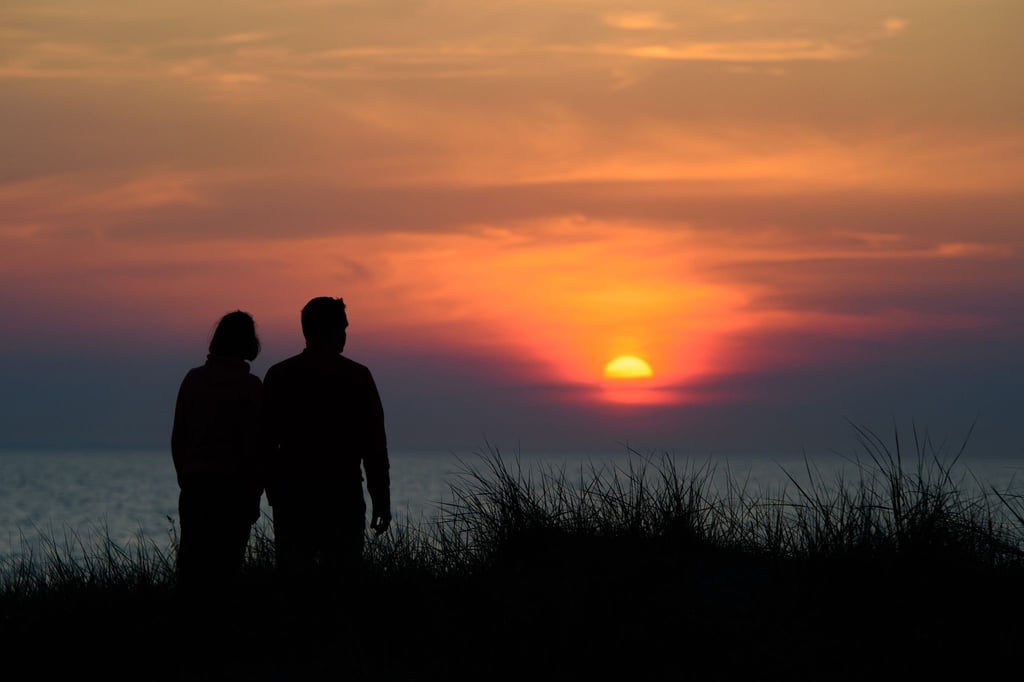 Ein Paar steht während des rötlich-orangenen Sonnenunterganges über der Ostsee an einer Düne am Strand.