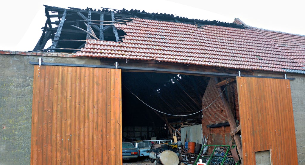 Der Morgen nach dem Feuer in Schenkenhorst. Der Brandgeruch liegt noch in der Luft, vor dem Vierseitenhof liegen Dachziegel und Stücke verbrannter Balken auf der Straße. Der Dachstuhl der großen Scheune, die die Anwohner als Lager nutzten, ist an einer Ecke völlig eingebrochen.