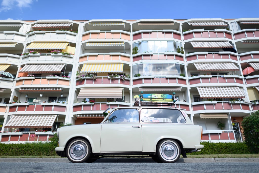 Ein Trabant Kombi 601s, der auf seinem Dachgepäckträger einen Koffer und ein Longboard geschnallt hat, steht vor einem Neubaublock.