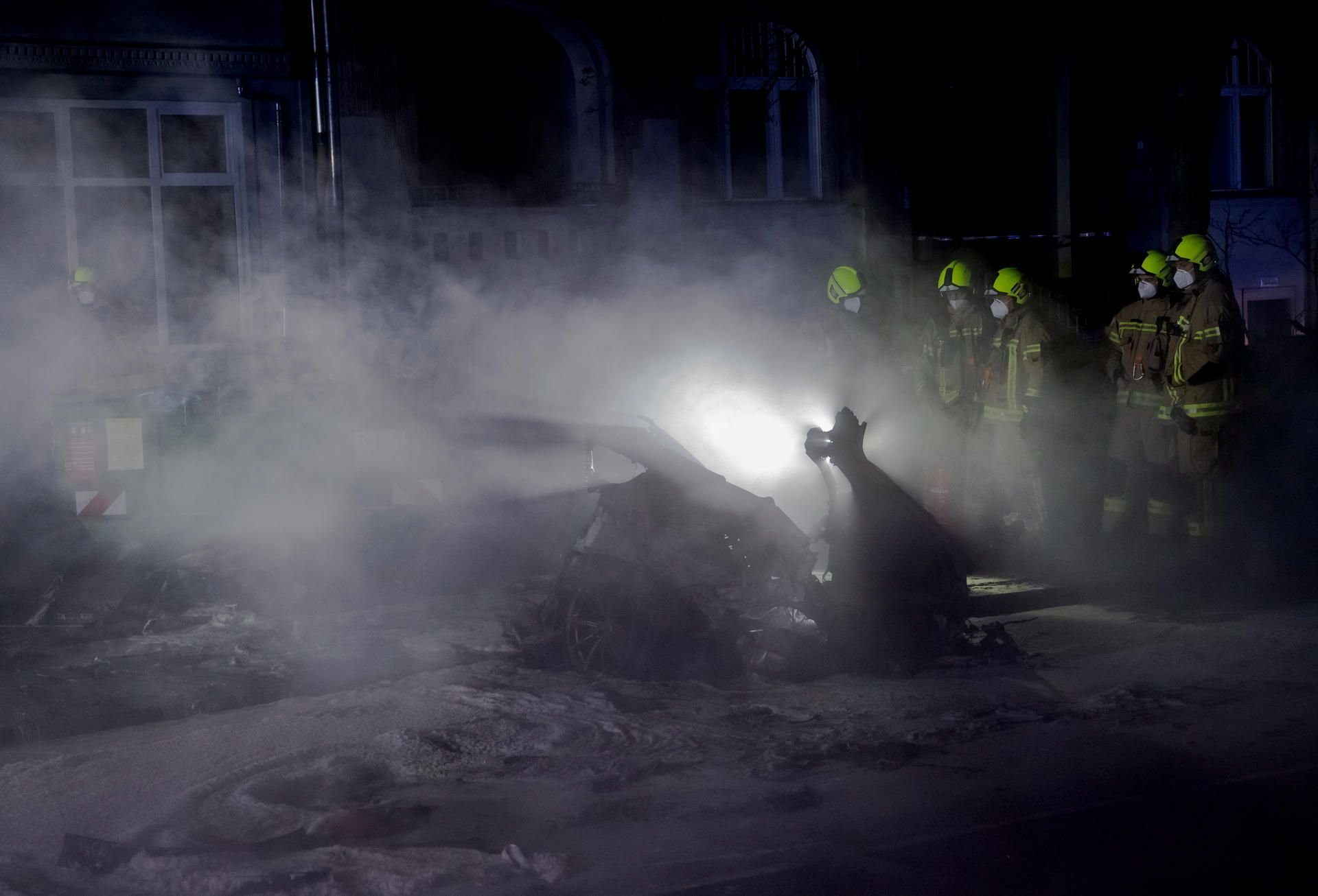Feuerwehrleute stehen am Treptower Park neben einem qualmenden Autowrack.
