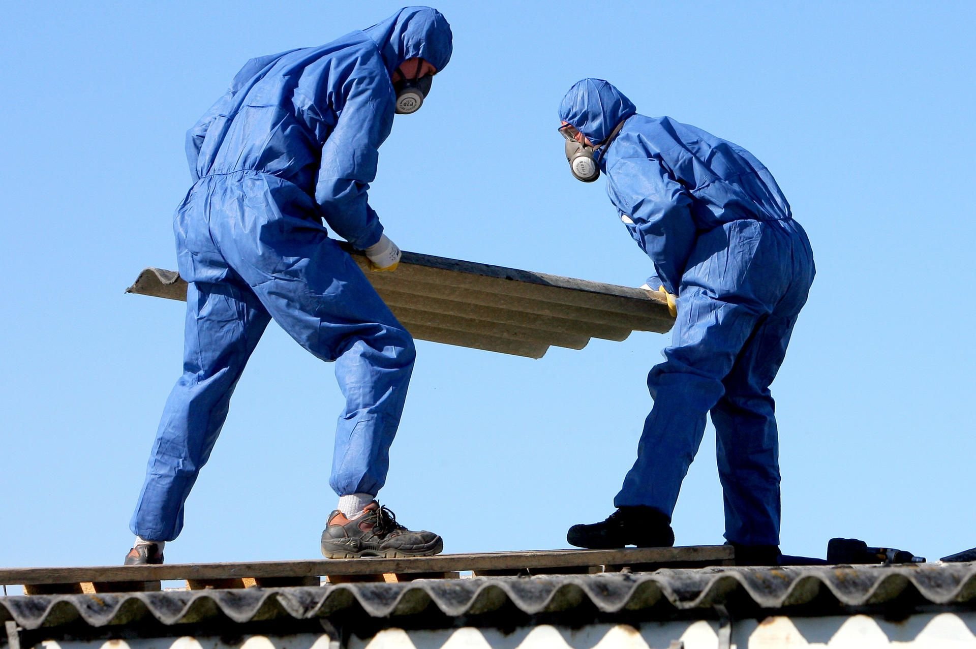 Nur etwas für Profis: Arbeiter in Schutzanzügen und mit Luftfiltern über Mund und Nase entfernen Asbestplatten vom Dach eines alten Gebäudes.