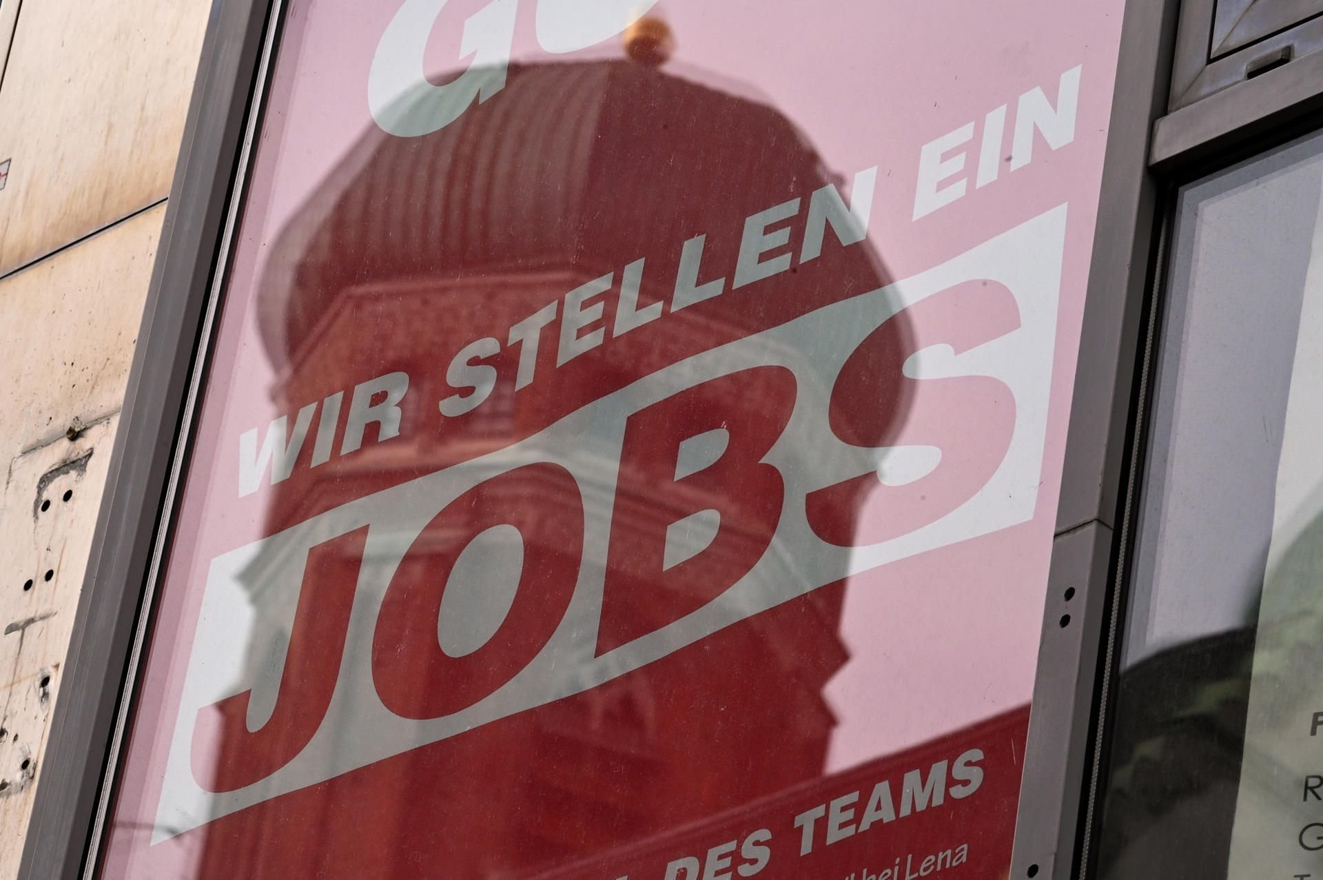 Ein Turm spiegelt sich in einer Fensterscheibe, in dem ein Schild mit der Aufschrift „Wir stellen ein - Jobs“ steht.