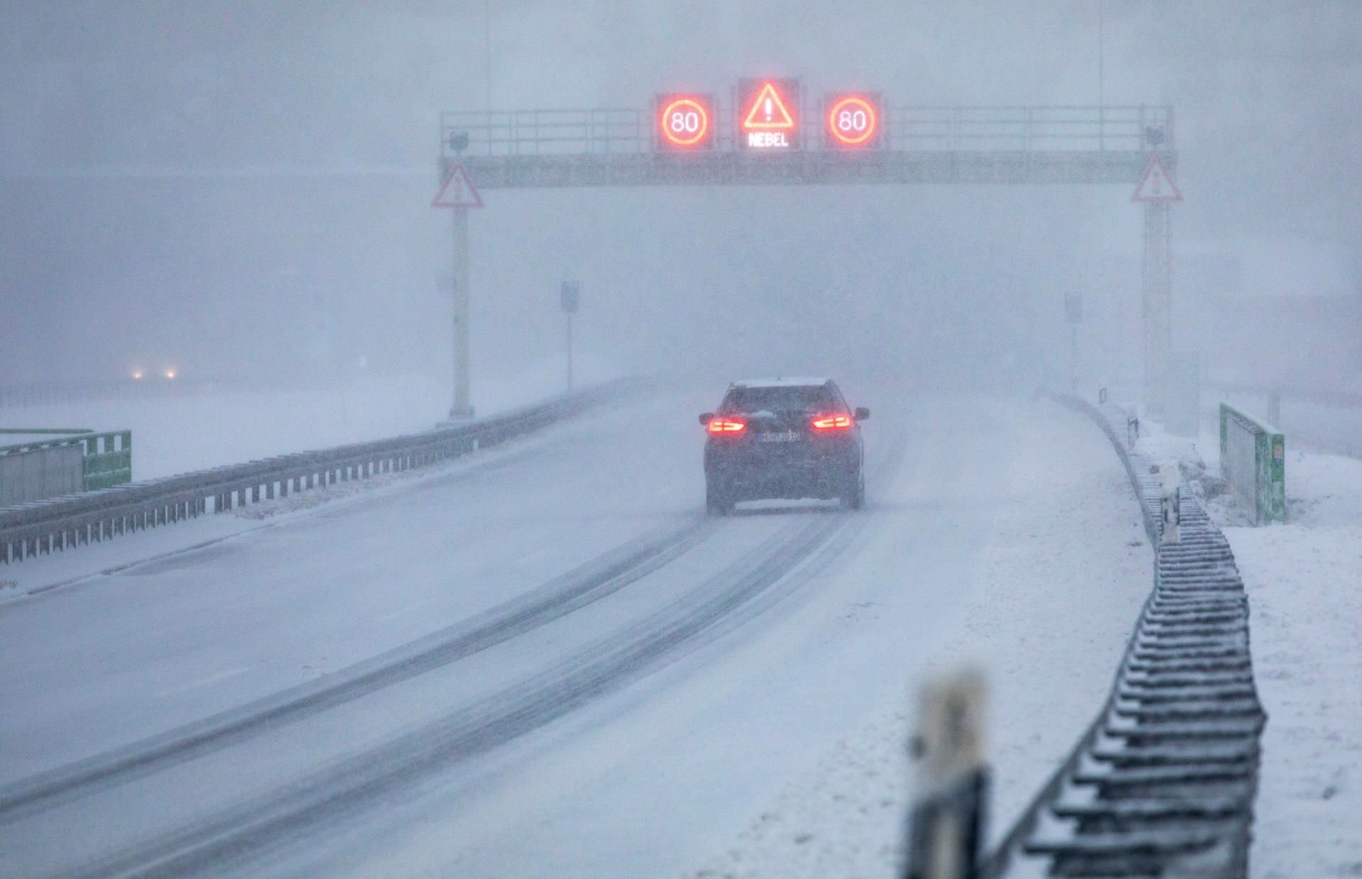 Thüringen, Zella-Mehlis: Ein Auto fährt auf der mit Schnee bedeckten A 71 unter einer Schilderbrücke durch, auf der die Verkehrszeichenanlage auf Nebel hinweist.