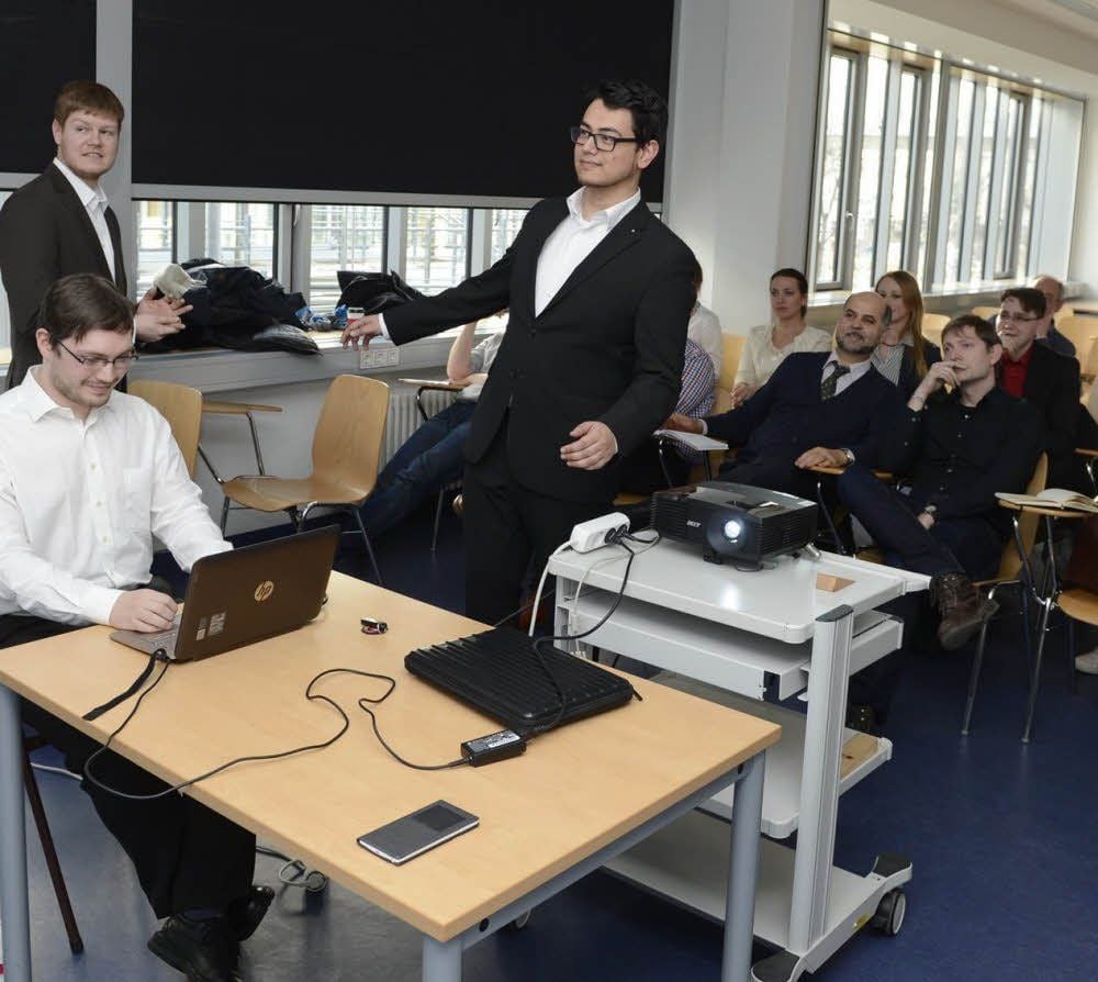 Andy Kämmer versucht, mittels Gewichtsverlagerung eine Kugel auf einem Spielbrett in ein Loch zu bewegen. Das Computerspiel wird an die Wand vor ihm projiziert, während Benjamin Körner (links stehend) erklärt.