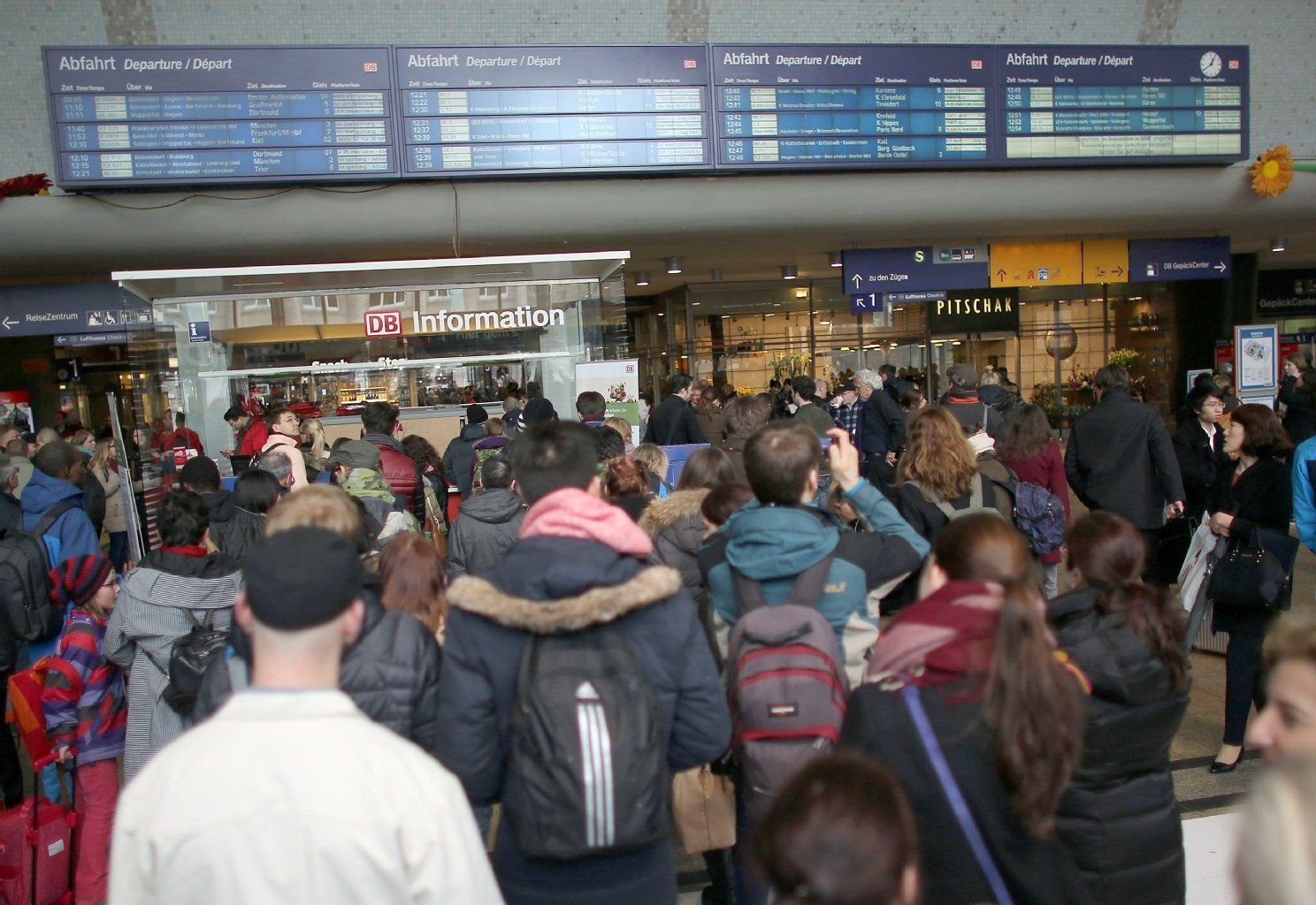 Bahnreisende stehen in Köln in einer Schlange an einem Informationsschalter am Hauptbahnhof während andere Reisende auf die Anzeigetafeln blicken.