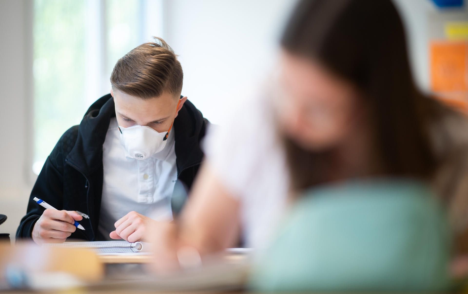 Schülerinnen und Schüler, einer davon mit einer Schutzmaske, bearbeiten Schulaufgaben.