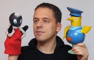 Mit dem Maskottchen der ehemaligen DDR-Tankstellenkette «Minol», dem «Minolpirol» (r.) und der beliebten Figur «Pittiplatsch» vom DDR-Kinderfernsehen steht der Sammler Eric Palitzsch in seiner Wohnung im sächsischen Freital. (FOTO: DPA)