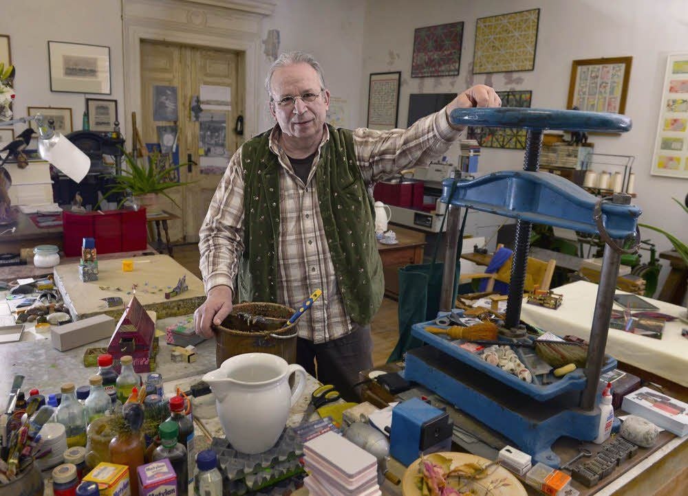 Papierkünstler Andreas Richter in seinem Atelier, in dem es viel zu entdecken gibt: Auf dem Arbeitstisch steht der Leim neben fertigen Schachteln, an der Wand hängen frühere Arbeiten im Rahmen - und zwischendrin finden sich historische Maschinen aus der Buchbinderei.