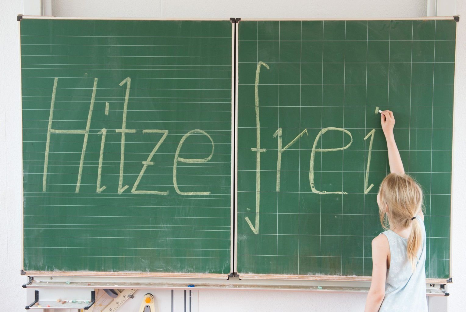 Eine Schülerin einer 2. Klasse schreibt in einer Grundschule das Wort "Hitzefrei" mit Kreide an eine Tafel.