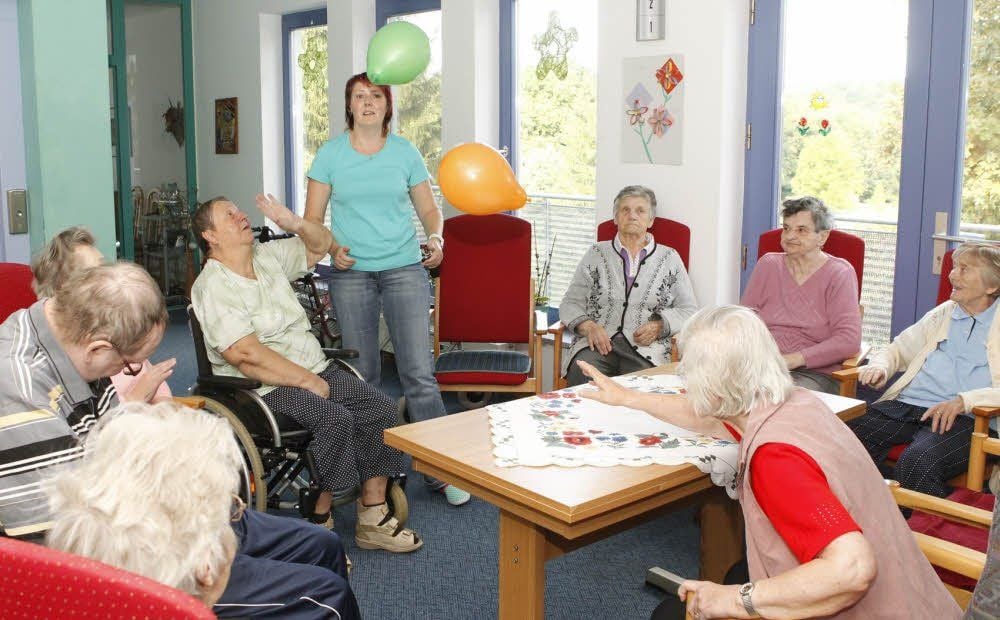 Auf dem Flur einer Etage des Altenpflegeheims macht eine Betreuerin mit Bewohnern Bewegungsübungen mit Luftballons.