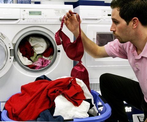 Ein Mann hockt vor einer Waschmaschine und überlegt, ob er den BH noch in die Maschine stecken soll. (FOTO: DPA)