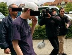 Ein junger Mann wird in Begleitung eines Polizeibeamten in Zivil (l.) und im Beisein eines Kamerateams am Dienstag (25.05.2004) in Hildesheim in des Landgericht geführt. Er war zum Prozessbeginn nicht erschienen. Der Jugendliche gehört zu den Angeklagten in einem Prozess, in dem eine monatelange Misshandlung eines Hildesheimer Berufsschülers verhandelt wird. (Foto: dpa)