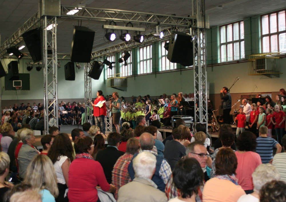 Die Turnhalle in der Schlossstraße von Annaburg zeigte sich am Freitagabend bestens gefüllt. Hier sprechen gerade Eva-Maria Glüer und Bianca Heinisch (auf der Bühne) die begrüßenden Worte ans Publikum, im Hintergrund die Generationen-Chöre und die ebenso altersgemischte Band.
