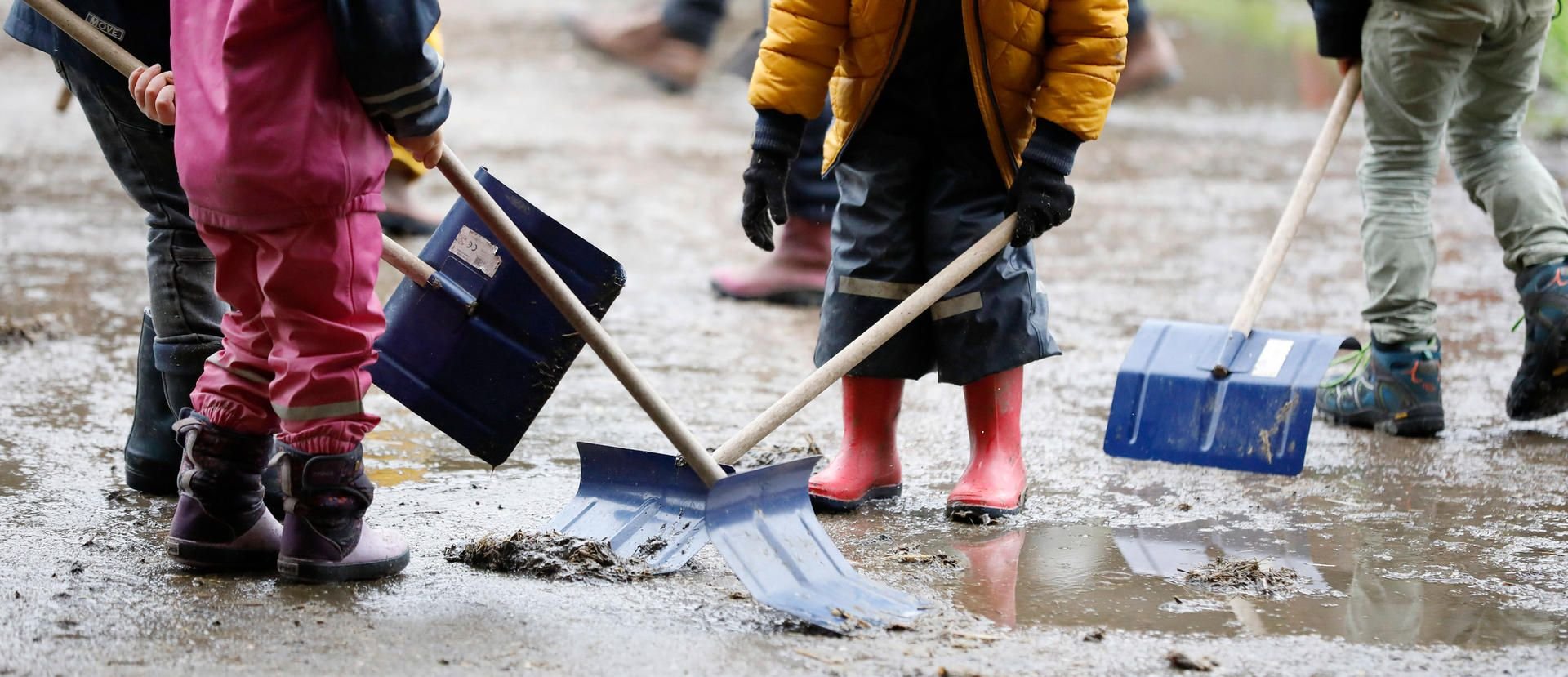 Kinder spielen mit Regenhosen und Schaufeln im Matsch.