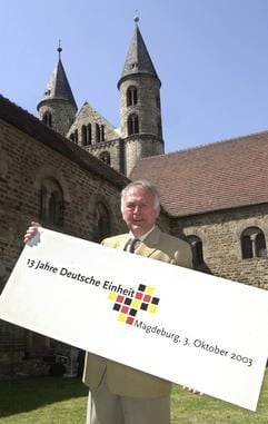 Sachsen-Anhalts Ministerpräsident Wolfgang Böhmer (CDU) posiert am Montag (04.08.2003) am Rande einer Pressekonferenz vor dem Kloster Unser Lieben Frauen in Magdeburg mit einem Schild, das auf die Feierlichkeiten zum Tag der Deutschen Einheit hinweist. (Foto: dpa)