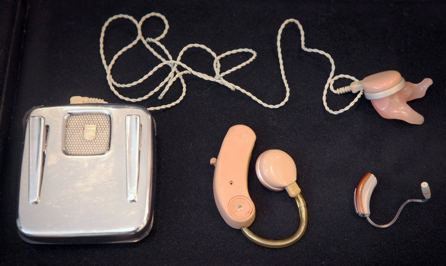 Drei Generationen von Hörgeräten, links ein altes Gerät aus den 1950ern, wo die Batterie-Kassette am Hemd befestigt werden musste und ein langes Kabel das Ohrstück verband, in der Mitte das erste kompaktere aus den frühen 1980ern und rechts unten ein modernes Hörgerät, aufgenommen am 27.02.2013 im Geschäft von Hörgeräteakustiker Bonsel in Mainz (Rheinland-Pfalz).