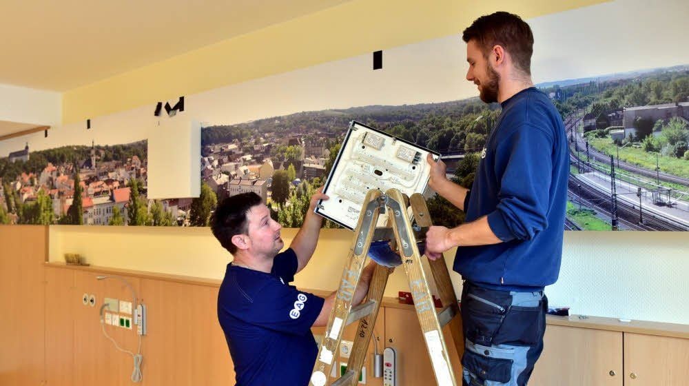 Michael Lämmchen und Christoph Nolze (v. li.) montieren in einem Patientenzimmer Lampen. Im Hintergrund die Fotowand, die die Region zeigt.