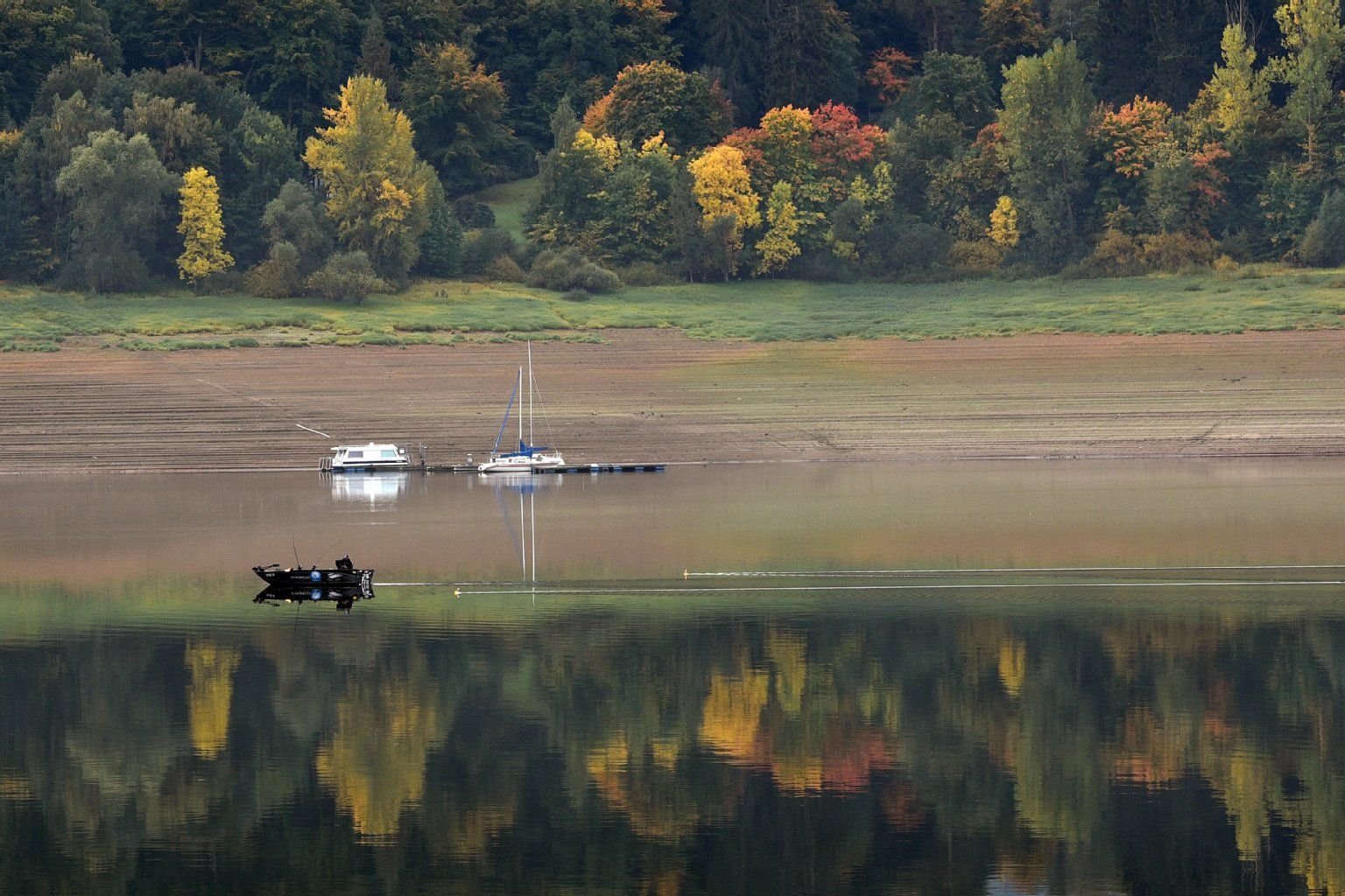 Ein Angler fährt mit seinem Boot auf dem Edersee an herbstlich verfärbten Laubbäumen vorbei, die sich auf der Wasseroberfläche spiegeln.