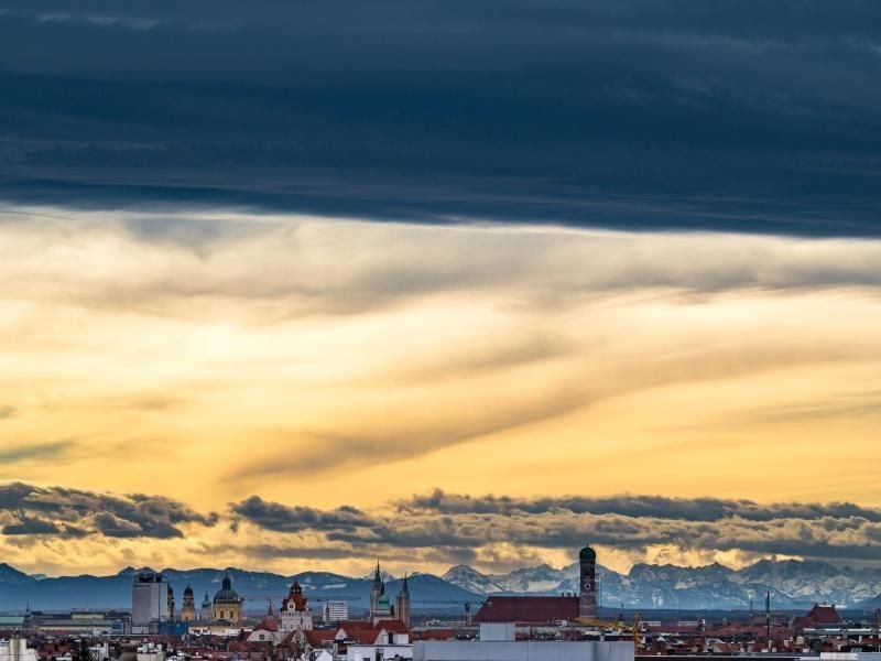 Ein dunkles Wolkenband hängt über der bayerischen Landeshauptstadt, deren Zentrum von den hohen Türmen der Frauenkirche (r.) markiert wird. Hinter der Skyline der Stadt ist das Alpenpanorama zu erkennen. Foto: Peter Kneffel/dpa