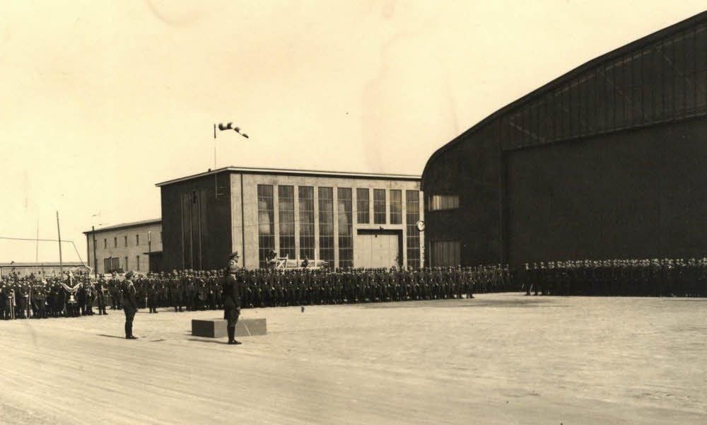 Die historische Aufnahme zeigt den Flugplatz mit Werfthalle, Werkstatt, Verwaltung, Kfz-Halle. Der Gruppenkommandeur Major Schwabedissen hält eine Ansprache. Alle Gebäude wurden Mitte der 1990er Jahre abgerissen.