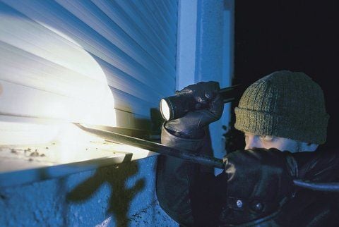 Ein Einbrecher mit Taschenlampe und Brechstange schaut durch die Lamellen einer Jalousie.