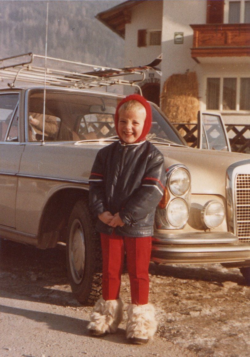 Winterurlaub 1973: Oliver trägt eine rote Mütze, ein rote Hose mit Bügelfalten und zottelige, weiße Fellschuhe. Hinter ihm glänzt ein Mercedes 280 SE in der Sonne.