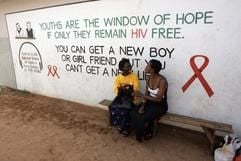 Zwei junge Frauen sitzen in Lusaka in Sambia auf einer Bank (Archivfoto). An der Wand hinter ihnen steht der Schriftzug einer Aids-Aufklärungskampagne. (Foto: dpa)