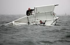 Besatzungsmitglieder eines gekenterten Bootes der olympischen Bootsklasse 49er versuchen am Mittwoch (23.06.2004) auf der Kieler Förde bei Kiel-Schilksee ihr Boot wieder aufzurichten. Wenig später wurde die Regatta auf Grund des schlechten Wetters abgebrochen. Das Wetter in den kommenden Tagen soll in Norddeutschland auch weiterhin regnerisch bleiben. Auf der Kieler Woche tragen noch bis Sonntag (27.6.2004) rund 6000 Segler aus 50 Nationen Regatten aus. (Foto: dpa)