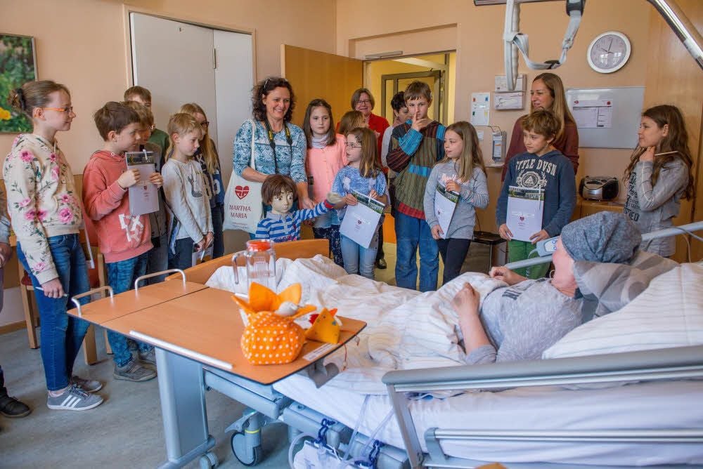 Viertklässler besuchen eine Patientin auf der Palliativstation. Auch die Puppe Jonas ist dabei. Sie steht am Fußende.