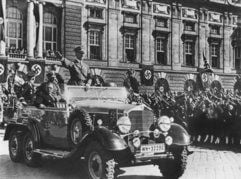 In einem Auto stehend grüßt Adolf Hitler die ihm zujubelnde Menschenmenge in Wien, neben ihm sitzt der österreichische Kanzler Arthur Seyss-Inquart (Archivfoto vom 14.03.1938). (Foto: dpa)