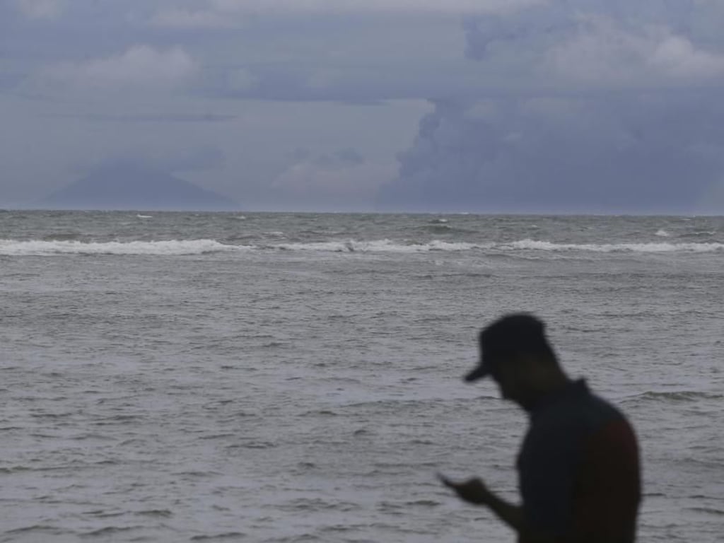 Ein Mann sieht in Carita, Indonesien, auf sein Handy. Im Hintergrund der Vulkan Anak Krakatau. Foto: Achmad Ibrahim/AP