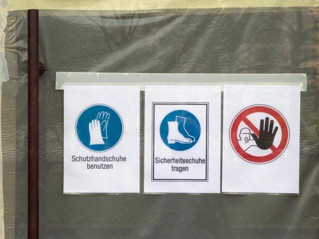 Schilder mit der Aufschrift für "Schutzhandschuhe benutzen", "Sicherheitsschuhe tragen" hängen an einer Baustelle. Foto: Soeren Stache/Archiv