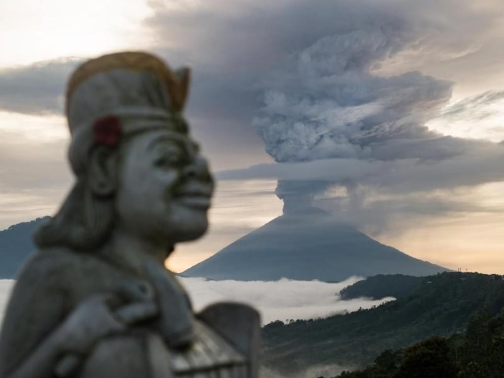 Entrücktes Lächeln: Eine Statue ist neben der riesigen Rauchsäule zu sehen, die über dem balinesischen Vulkan Mount Agung aufsteigt. Foto: Josh Edelson