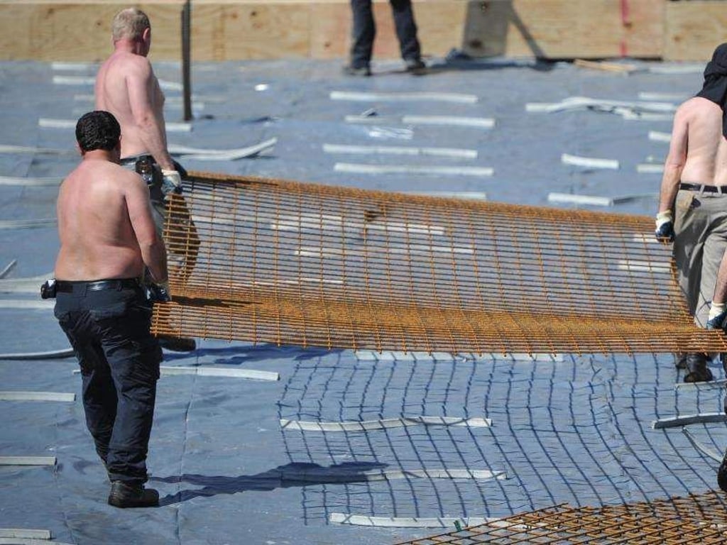 Arbeiter mit freiem Oberkörper tragen auf einer Baustelle ein Stahlgitter. Foto: Ole Spata