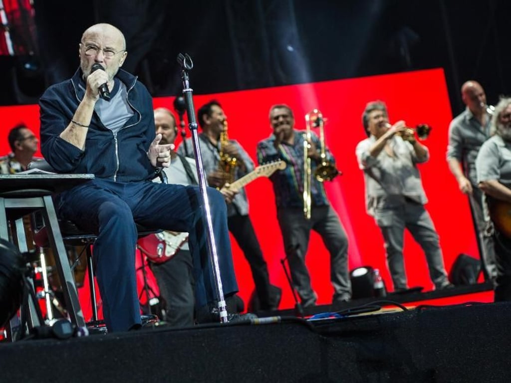 Sänger Phil Collins sitzt auf der Bühne und singt, hinter ihm stehen die Bandmitglieder. Foto: Christoph Schmidt