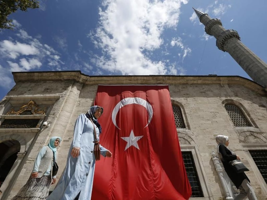 Frauen gehen in Istanbul an einer Moschee vorbei, an der eine große türkische Fahne hängt. Die USA wollen nicht lockerlassen, bis der in der Türkei in Hausarrest sitzende US-Pastor Andrew Brunson wieder bei seiner Familie sei. Foto: Lefteris Pitarakis/AP