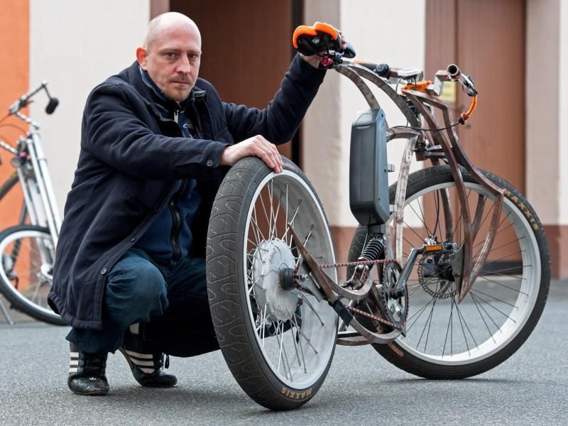Christian Kelemen hockt vor seiner Fahrrad-Werkstatt im Fischereihafen von Bremerhaven (Bremen) neben seinem E-Bike im Chopper-Stil mit besonders hoch positionierten Sattel. Foto: Ingo Wagner