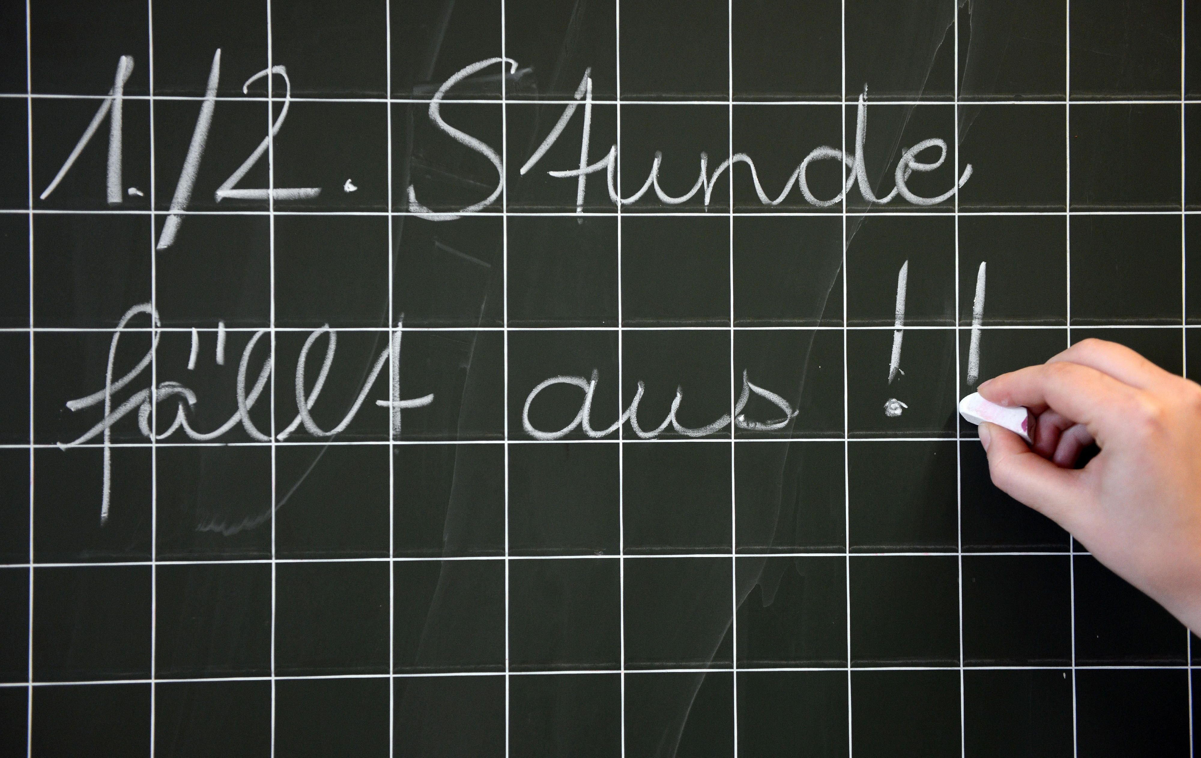 Eine Frau schreibt in einer Schule „1./2. Stunde fällt aus!!“ an die Tafel.