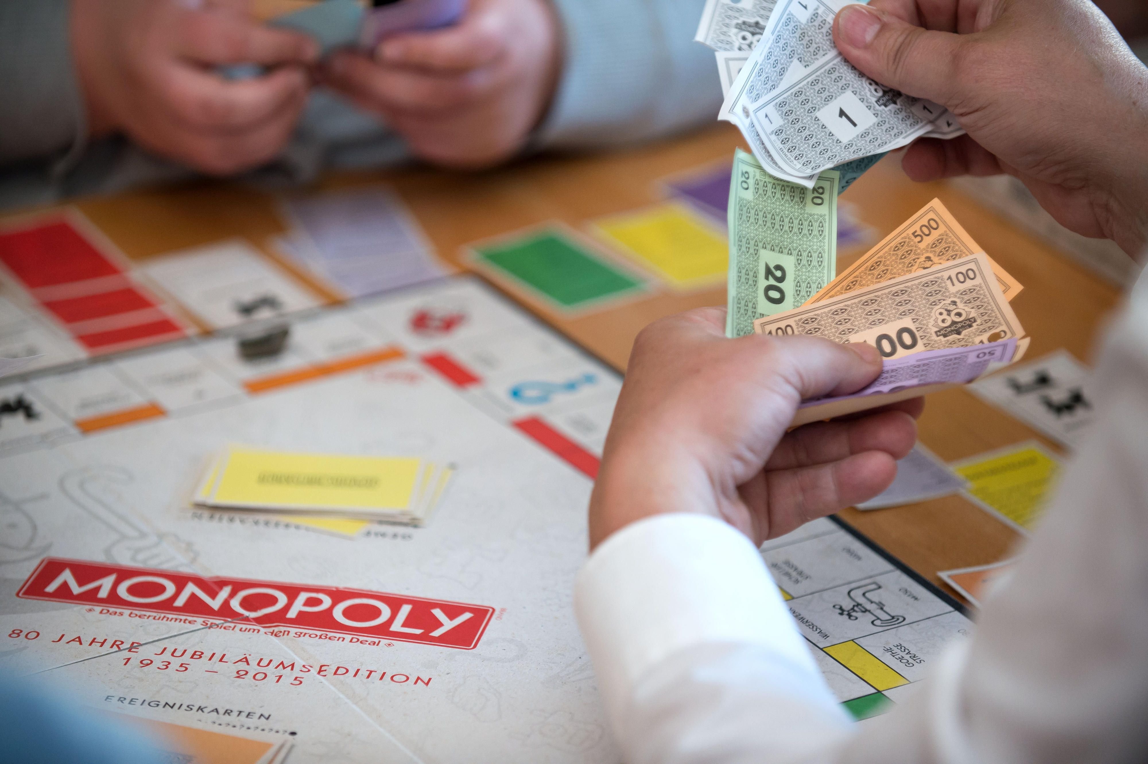 Spieler sitzen vor einem Monopoly-Spiel und halten Spielgeld in der Hand.