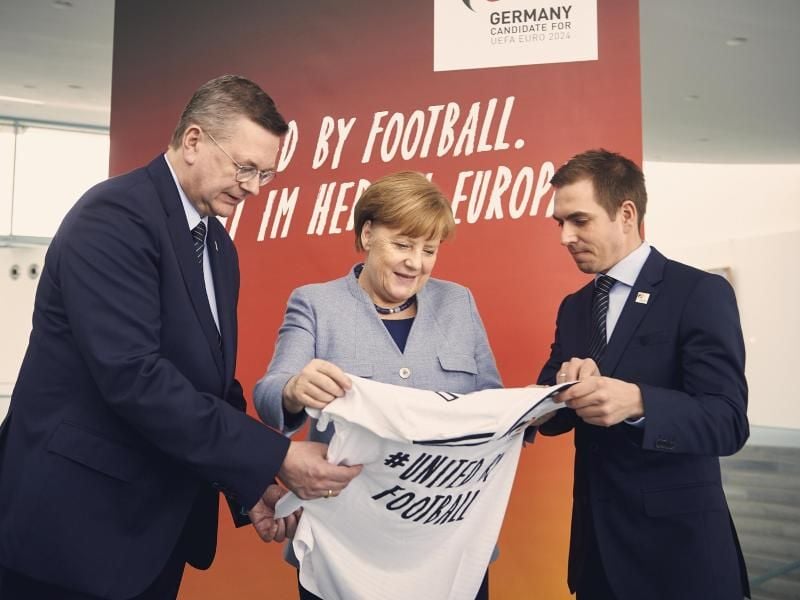Bundeskanzlerin Angela Merkel (M) bekommt anlässlich der Bewerbung für die Fußball-EM 2024 von DFB-Präsident Reinhard Grindel (l) und EM-Botschafter Philipp Lahm ein Trikot überreicht. Foto: Felix Zahn/Bundesregierung